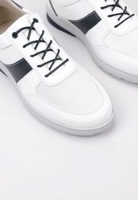 Chaussures blanches en mesh et synthétique avec des accents noirs, à lacets, à bout arrondi et semelles en caoutchouc. La forme générale est sportive et décontractée.