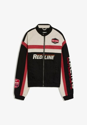 Junger Mann trägt eine rot-weiß-schwarze Jacke, weißes Hemd, schwarze Shorts und Sandalen und steht an einer geschwungenen weißen Wand drinnen gelehnt.