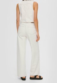Weißer Jeans-Overall mit weitem Bein und ärmellosem Design. Verfügt über Gesäßtaschen und kontrastierende Nähte. Das Model trägt schwarze Sandalen.