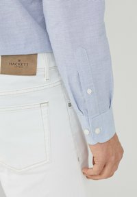 Camicia a maniche lunghe a righe blu chiaro con polsini bianchi, abbinata a jeans bianchi. Etichetta Hackett London visibile sulla cintura.