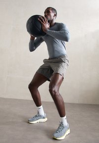 Mand i grå sportstop og shorts holder en sort medicinbold og udfører en squat i et minimalistisk indendørs rum.