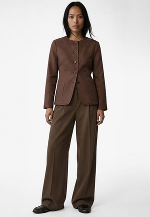 Kvinde stående iført brun knapjakke, bredbenede brune bukser og mørkebrune sko mod en ensfarvet baggrund.