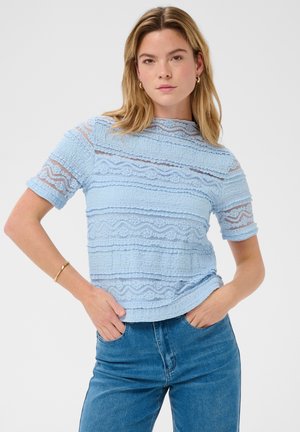 Jeune femme aux cheveux blonds portant un haut en dentelle bleu clair et un jean bleu, debout les mains dans les poches sur fond blanc.