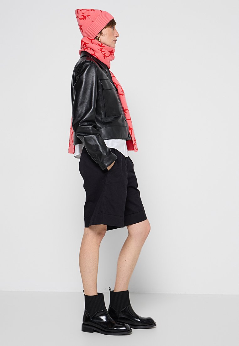 Schwarze Lederjacke, pinkfarbene, gemusterte Schal, hellpinkfarbene Mütze, schwarze Shorts und glänzende schwarze Stiefeletten, die einen modernen, lässigen Outdoor-Look präsentieren.