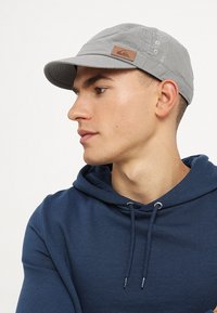 Gorra gris en mezcla de algodón, con una corona redondeada, visera corta y rígida, y un parche de logo de cuero marrón en el lateral.