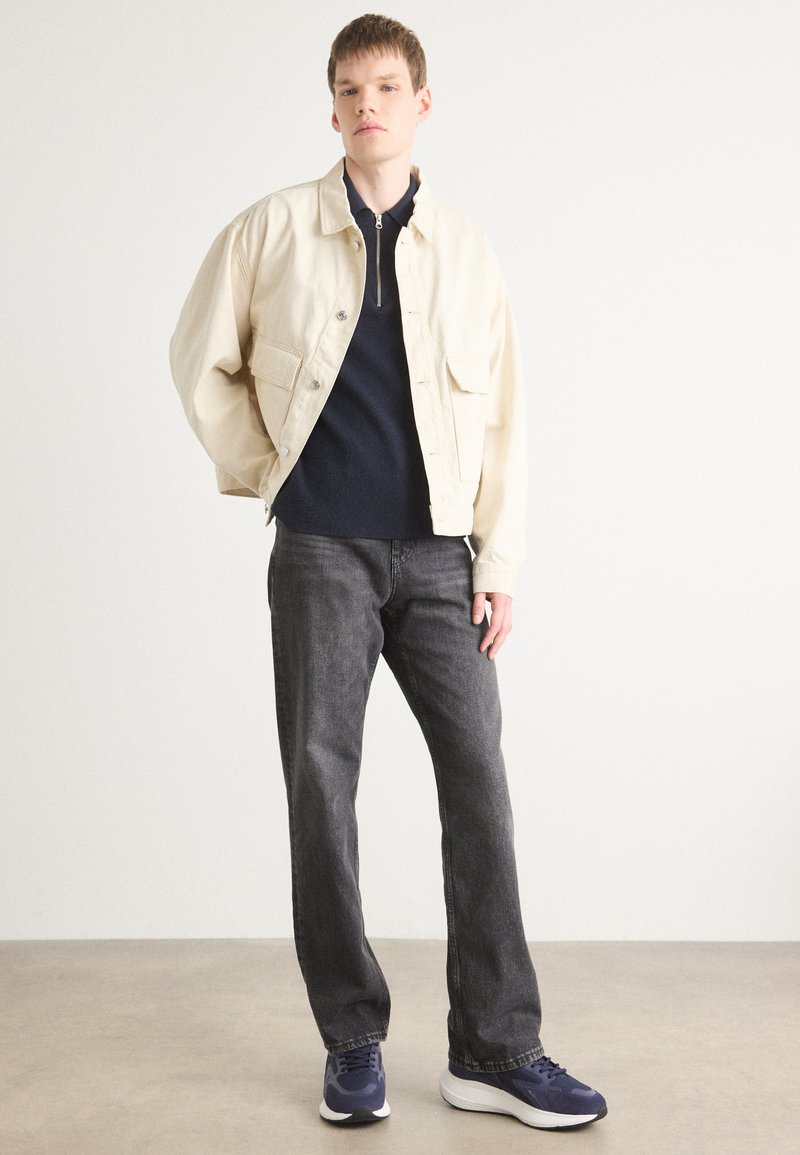 Crème-kleurige jas met twee voorzakken, marineblauwe trui eronder, donkergrijze jeans en marineblauwe sneakers. Textuurstof, relaxte pasvorm.