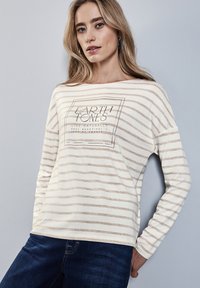 Cremfarbener Langarmoberteil mit hellbraunen Streifen, versehen mit einem quadratischen Grafikdesign, das "Erdtöne" liest, und lockerer Passform. Kombiniert mit dunkelblauen Jeans.