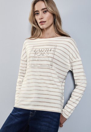 Haut créme à manches longues avec des rayures brun clair, mettant en avant un graphique carré portant l'inscription "Earth Tones" et une coupe décontractée. Porté avec un jean bleu foncé.