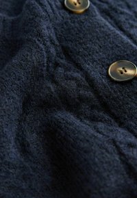 Mornarsko modra pletena oblačila iz teksturirane tkanine, ki ima dva okrogla, rjava gumba z štirimi luknjami vsak. Vidna šivna vzorec.