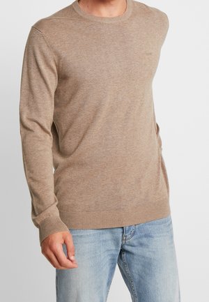 Mand iført en beige langærmet rundhalset sweater med ribmønstrede ærmegab og kant kombineret med lyseblå jeans.