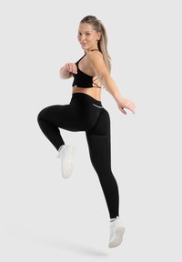 Top corto sportivo nero con design a incrocio sulla schiena, abbinato a leggings neri a vita alta. Modella che indossa scarpe da ginnastica bianche.