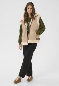Beige mouwloze vest met crème nepbont en ritssluiting. Gemaakt over een groene sweatshirt en zwarte broek, aangevuld met bruine schoenen.