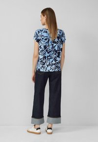 Blauwe bloemenblouse met korte mouwen en donkerblauwe patronen, gecombineerd met donkere wijde jeans met opgerolde broekspijpen en witte sneakers.