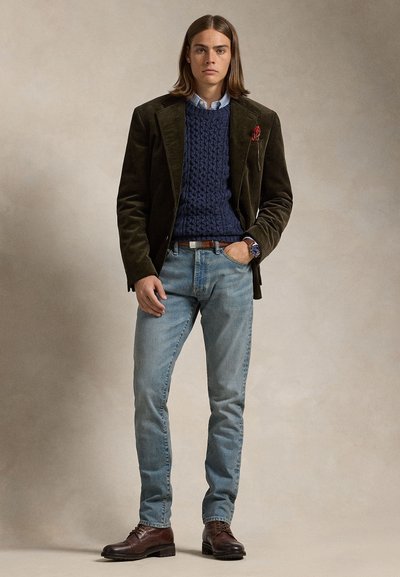 Mladý člověk nosící tmavě hnědý manšestrový blejzr, námořnicky modrý svetr s copánkovým vzorem, světle modré džíny a hnědé kožené boty, stojící na neutrálním pozadí.