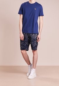 Granatowa bawełniana koszulka z okrągłym dekoltem i małym haftowanym logo; granatowe spodenki w wzór z białymi plamami; białe sneakersy.