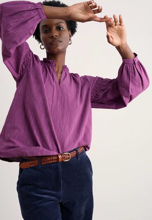Paarse gestreepte blouse met volumineuze mouwen, een mandarincol en gathered details. Draag met donkere corduroy broek en een gevlochten bruine riem.
