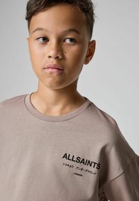 Gaiši brūns īsās piedurknes T-krekls ar apaļu kaklu. Melna teksta grafika kreisajā pusē ar "ALLSAINTS" un japāņu rakstzīmēm.