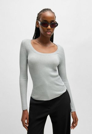 Hellgraues langärmeliges Rippoberteil mit weitem Ausschnitt, figurbetonter Passform, kombiniert mit schwarzen Hosen und kantigen, dunklen Sonnenbrillen.