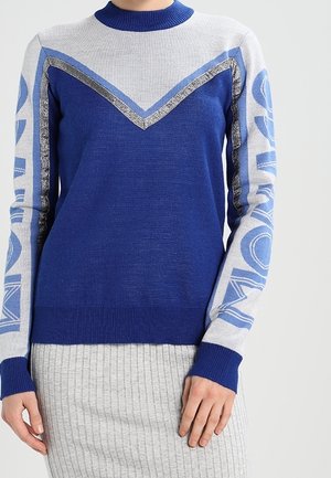 Blå og lysegrå sweater med chevronmønster og fed skrift på ærmerne, båret med en ribbet grå nederdel.