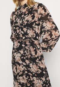 Czarna sukienka w kwiaty z wysokim kołnierzem, długimi, bufkowymi rękawami i przewiązanym w pasie. Kwiatowe wzory w odcieniach różu i beżu tworzą strukturalny wygląd.