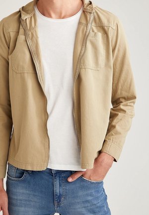 Uomo con giacca beige con zip sopra una maglietta bianca e jeans blu, con la mano destra in tasca, su uno sfondo chiaro e semplice.