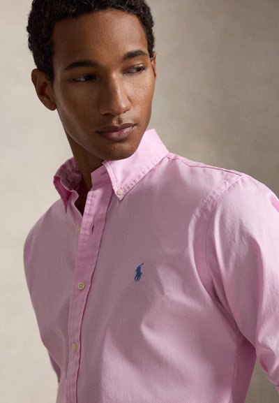 Άντρας που φοράει ανοιχτό ροζ πουκάμισο με κουμπιά και μπλε κεντημένο λογότυπο, κοιτάζοντας αριστερά σε ουδέτερο φόντο.