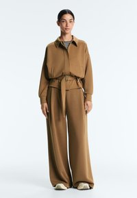 Žena stojaca v hnedom páse s opaskom a zodpovedajúcich širokých nohaviciach, cez čierne a biele pruhované tričko a béžové tenisky.