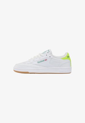 Zapatilla baja blanca de Reebok con logo verde, detalle de la bandera Union Jack y lengüeta del talón en amarillo neón, con suela de goma y cordones blancos.