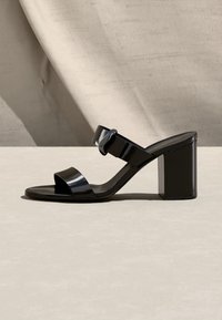 Μαύρο σανδάλι με τακούνι από δερμάτινο λουστρίνι με δύο λουριά, το ένα με αγκράφα, και χοντρό τακούνι τύπου block σε ανοιχτή επιφάνεια.