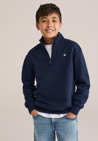 Temno modra zip-up dukserica z visokim ovratnikom in rebrastimi manšetami, z logotipom na prsih in oblečena nad belim majico.