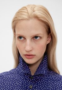 Navy blue shirt with white polka dots, button-up design, and high collar. Smooth texture, features visible buttons and stitching details.