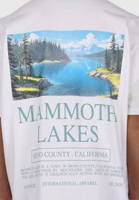 White t-shirt featuring a scenic printed design of a lake with trees and mountains, along with "MAMMOTH LAKES" text in light blue.