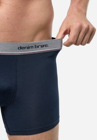 Marineblaue Boxershorts aus weichem Stoff, mit einem breiten grauen Bund, der ein Markenlogo und kontrastierende Streifen zeigt.