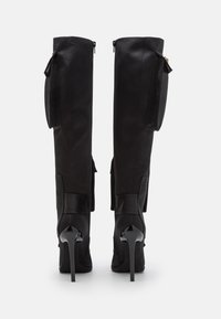 Bottes noires montantes jusqu'à la cuisse en cuir lisse, talons pointus et brillants, fermeture éclair à l'arrière, et poches latérales pour plus de détails.