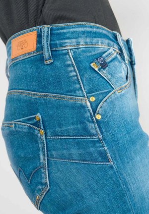 Jean en denim bleu au design ajusté, avec une étiquette en cuir marron, des coutures jaunes et des poches supplémentaires avec des boutons dorés.