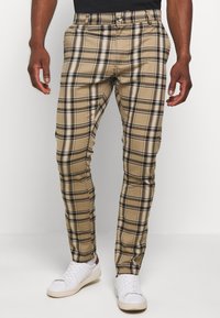 Beige plaid trousers made of cotton with black and white accents, featuring a fitted design and front pockets. Worn with white sneakers.