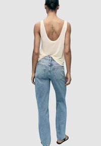 Top senza maniche color beige chiaro con scollatura profonda sul retro, abbinato a jeans azzurri a vita alta e sandali neri. Design semplice e vestibilità rilassata.