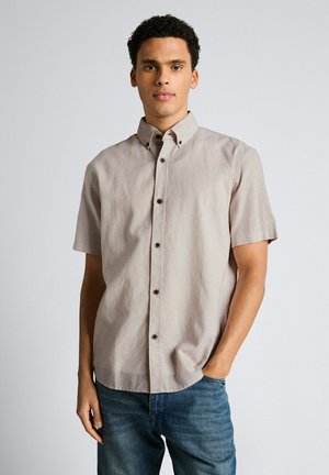 Mann trägt hellbeiges kurzärmeliges Hemd mit Knöpfen und blaue Jeans, steht mit einer Hand in der Tasche vor einfarbigem Hintergrund.