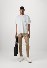 Hvit bomull t-skjorte med rødt logo, beige bukser, hvite joggesko og svart kviltet veske. Modellen har på seg en svart caps og står på en nøytral bakgrunn.