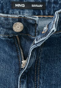Jean en denim bleu avec fermeture éclair partiellement ouverte et bouton en métal étiqueté MNG, étiquette visible indiquant la taille et fabriqué au Pakistan.