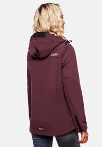 Woman with blonde wavy hair wearing a maroon hooded jacket with black and white logo patches, standing against a plain light background.