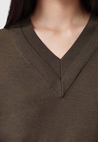 Sweter w kolorze oliwkowej zieleni z dekoltem w serek, wykonany z miękkiego materiału o gładkiej fakturze i widocznym przeszyciu wzdłuż linii dekoltu.