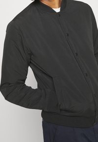 Veste bomber noire avec un col et des poignets côtelés, dotée de boutons-pression et de poches latérales, fabriquée en tissu lisse et léger.