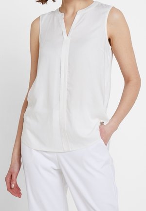 Nainen pukeutuneena valkoiseen hihattomaan V-kaula-aukkoiseen paitaan ja valkoisiin housuihin, seisoo käsi taskussa valkoista taustaa vasten.