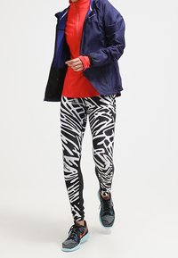 Juoksuasu, johon kuuluu punainen pitkähihainen paita, tummansininen takki ja mustavalkoiset abstraktikuvioiset leggingsit. Mukana siniset urheilukengät.