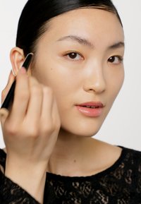 Makeupapplikator i handen, modellen applicerar produkt på sin kind. Hon har slät hud, mörkt hår och bär en svart spetsblus. Neutral bakgrund.