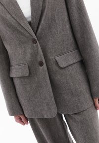 Harmaa raidallinen puku, jossa on viistetty kaulus, kaksi etutaskua ja tummat napit. Kankaan pinnassa on kudottu rakenne, jossa on hienovarainen kiilto.