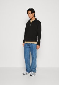 Sort langærmet trøje med lynlås op til brystet lagd over en beige skjorte, parret med lyseblå denimjeans og sølvfarvede sportsko.