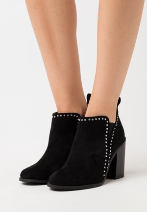 Černé kotníkové boty z broušené kůže s kulatou špičkou, zdobené stříbrnými cvočky podél horního okraje a masivním blokovým podpatkem. Měkká textura a stylový silueta.