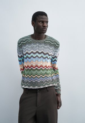 Jeune homme portant un pull rayé ondulé multicolore et un pantalon marron foncé, debout devant un fond gris clair uni.